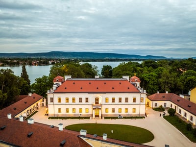 Herrenhaus mit rotem Dach am See, umgeben von Bäumen.