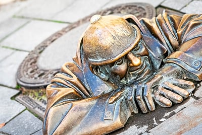 Bronzestatue eines Mannes, der aus einem Kanaldeckel schaut.