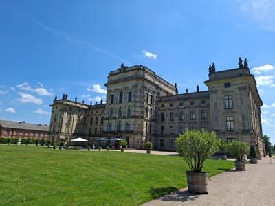 Historisches Gebäude mit Rasenfläche und Bäumen im Vordergrund unter blauem Himmel.