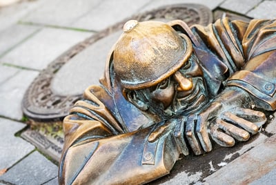 Bronzestatue eines Mannes, der aus einem Kanaldeckel schaut.