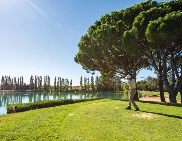 Golfer schwingt Schläger auf Platz mit Bäumen und See.